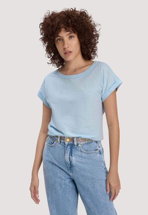 Frau mit lockigem Haar, die ein hellblaues T-Shirt mit hochgekrempelten Ärmeln trägt, in das eine hochtaillierte hellblaue Jeans mit einem gemusterten Gürtel gesteckt ist.
