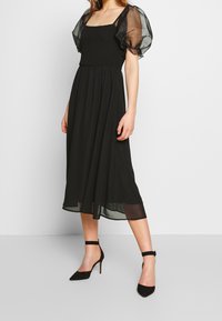 Schwarzes Kleid mit Puffärmeln und einem figurbetonenden, gerafften Oberteil. Der fließende Rock reicht bis zur Mitte der Wade. Getragen mit schwarzen Knöchelriemen-Absätzen. Glatte Textur.
