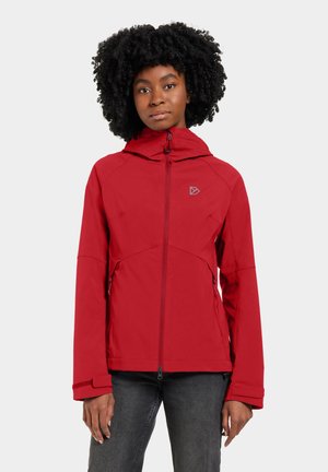 Frau mit lockigem Haar, die eine rote Jacke mit Reißverschluss und Kapuze sowie dunkle Hosen trägt, steht vor einem schlichten hellen Hintergrund.