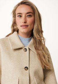 Světlý béžový kabát z vlny s příměsí, který má límec, dvě přední kapsy a dva hnědé knoflíky. Nosený přes světle modrý svetr.
