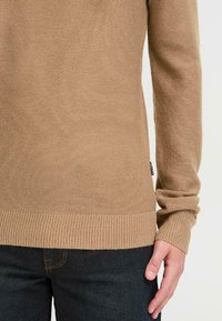 Jersey de punto beige con puños y dobladillo acanalados, mangas largas y un acabado texturizado. Combinado con jeans oscuros.