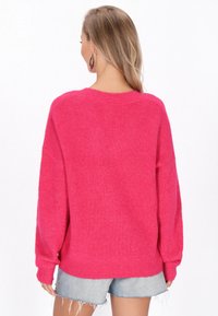 Pulover supradimensionat de un roz aprins, cu un tricot texturat, cu model pe față. Prezintă un decolteu larg și umeri lăsați, asortat cu șorturi din denim uzate.