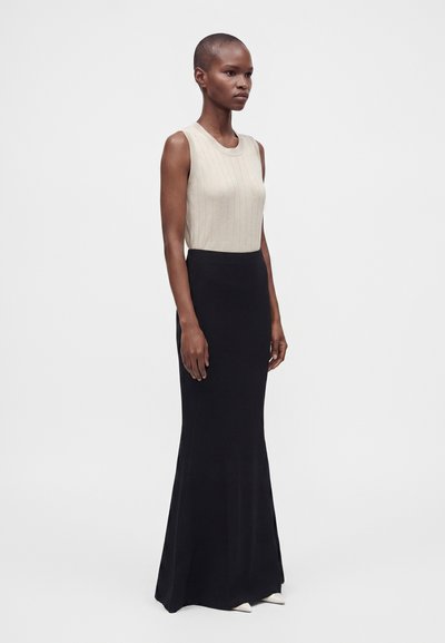 Woman with short hair wearing a sleeveless beige top and a long black skirt standing against a plain white background.