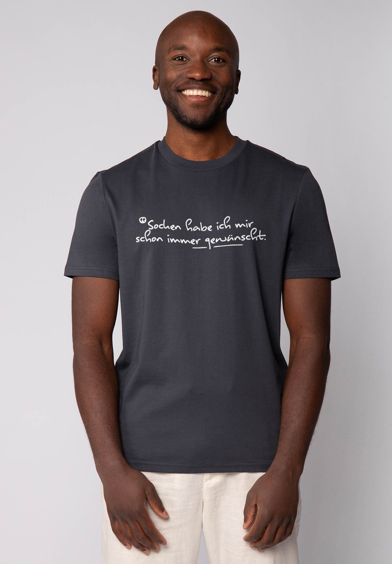 Glunderende man in een zwart T-shirt met witte Duitse tekst "Sochen habe ich mir schon immer gewünscht" en beige broek tegen een effen achtergrond.