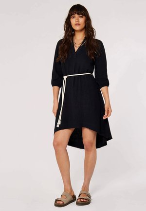 Loses schwarzes Kleid mit Dreiviertelärmeln, einem V-Ausschnitt und einem fransigen Saum. Gestylt mit einem weißen gebundenen Gürtel und getragen mit beigen Sandalen.