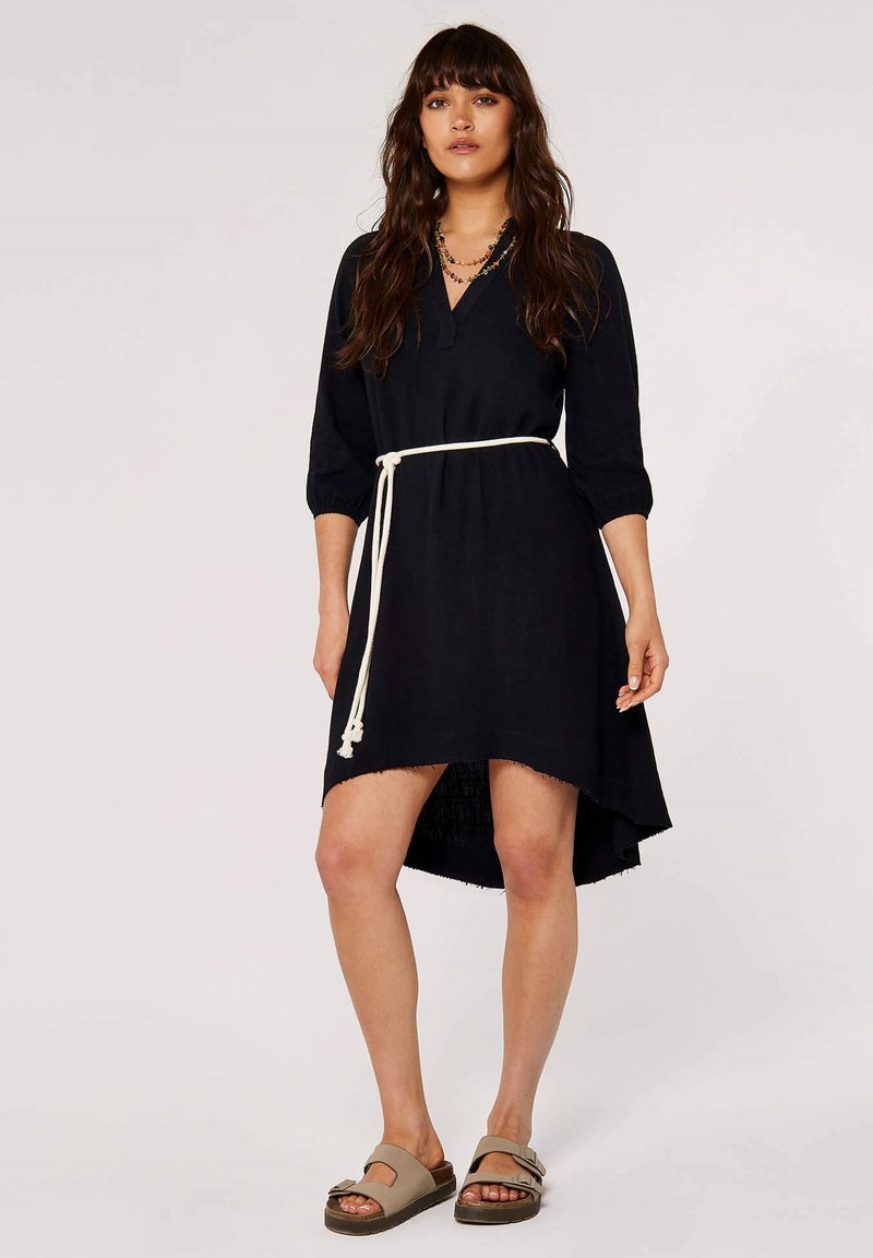 Loses schwarzes Kleid mit Dreiviertelärmeln, einem V-Ausschnitt und einem fransigen Saum. Gestylt mit einem weißen gebundenen Gürtel und getragen mit beigen Sandalen.