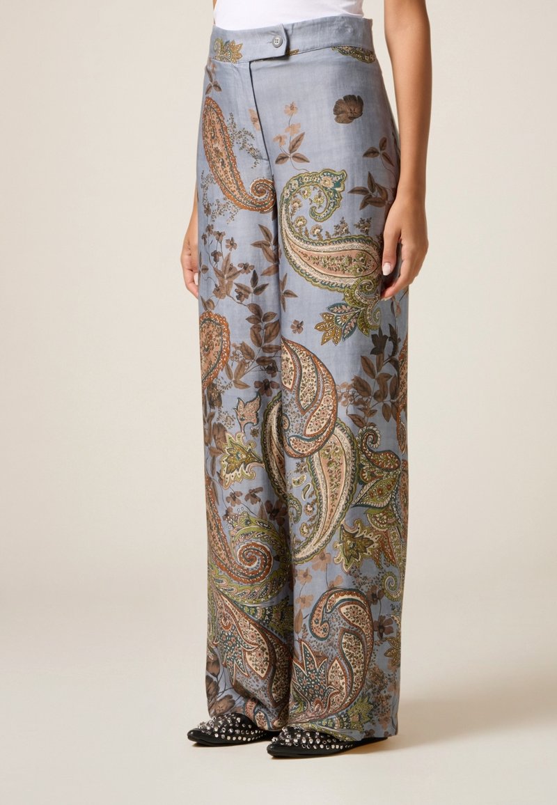 Femme portant un pantalon taille haute à jambes larges avec une base bleu et des motifs cachemire et floraux multicolores, assorti à des chaussures noires pointues cloutées.