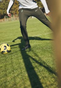 Zwarte sportieve schoenen, olijfgroene Nike-broek met Spurs-logo, witte longsleeve top, speler gepositioneerd naast een gele voetbal op gras.