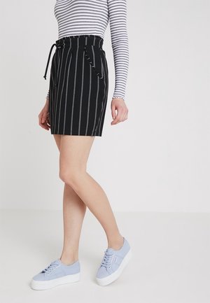 Personne portant une jupe noire taille haute à rayures verticales blanches, un haut rayé blanc et noir à manches longues, et des baskets compensées bleu clair.