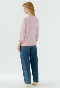 Femme vue de profil portant un pull duveteux rose clair, un jean bleu ample et des chaussures à talons beiges sur un fond clair uni.