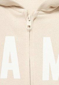 Felpa con zip beige realizzata in un tessuto texture, caratterizzata da grandi scritte bianche e una zip chiara. Design semplice e minimal.