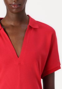 Polo-shirt rouge avec un col en V, manches courtes, tissu texturé et un petit logo sur la manche. Design épuré et ajusté.