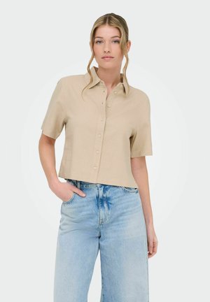 Młoda kobieta z rozpuszczonym blond włosami, ubrana w beżową koszulę na guziki z krótkim rękawem i jasnoniebieskie spodnie z wysokim stanem, z ręką w kieszeni.