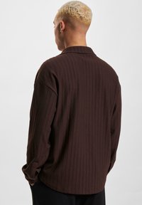 Chemise marron à côtes avec une coupe décontractée, dotée d'un col et de manches longues. Une texture remarquable et de fines lignes verticales mettent en valeur le design.