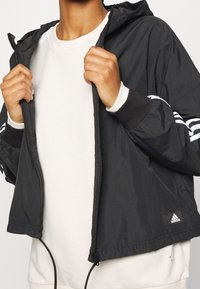 Giacca nera con zip realizzata in materiale leggero, dotata di cappuccio, accenti a strisce bianche sulle maniche e polsini elasticizzati.