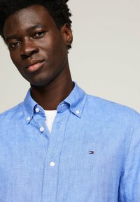 Camicia di lino azzurro chiaro con colletto button-down, bottoni bianchi e una piccola toppa con logo che presenta strisce blu navy, rosse e bianche.