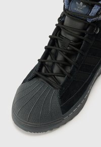 Sneaker en suède noir et cuir avec un bout en caoutchouc texturé, des lacets ton sur ton et des accents bleus, présentant trois rayures sur le côté et un design à coupe basse.