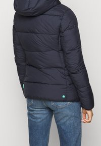 Doudoune marine avec capuche, présentant des sections matelassées horizontales, des poches latérales et un petit accent de logo vert près de l'ourlet.