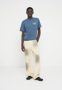 Μπλε μπλουζάκι με κίτρινο λογότυπο, συνδυασμένο με εκρού παντελόνι με μαύρο διαβαθμισμένο χρώμα στο τελείωμα. Μαύρα slip-on παπούτσια ολοκληρώνουν το σύνολο.