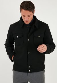 Juodas švarkas iš tekstūruotos medžiagos, su užsegamu priekiu ant sagų, dviem krūtinės kišenėmis ir klasikiniu apykakle. Vidus lygus.