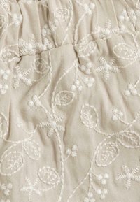 Tissu beige avec broderies florales blanches et motifs de feuilles. Surface texturée avec des accents en relief et des détails froncés.
