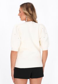 Hvid strikket sweater med korte puffærmer, der har pynt på skuldrene og en ribbet kant. Stylet med sorte shorts.
