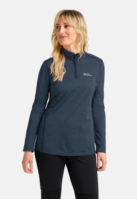 Blonde Frau, die einen navy-blauen Langarm-Quarter-Zip-Pullover und schwarze Hosen trägt, nach links schaut und leicht lächelt.