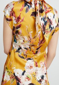 Robe en satin dorée avec un motif floral. Présente un col haut avec un nœud noué et des manches courtes, mettant en valeur des couleurs vives et une texture douce.