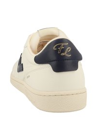 Sneaker in pelle bianca con suola testurizzata, inserto sul tallone blu navy con iniziali dorate "FL" e dettagli laterali, visto dall'angolo posteriore destro.