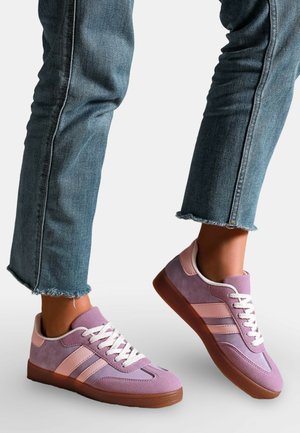 Jambes en jean à ourlet effiloché bleu portant des baskets violettes et roses avec des lacets blancs et des semelles brunes sur fond blanc.