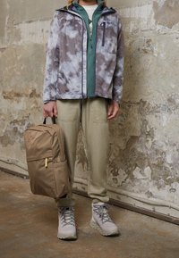 Lichte, waterdichte jas in het grijs met witte patronen, gelaagd over een groene ritssluiting, gecombineerd met khaki broek en een tan rugzak.