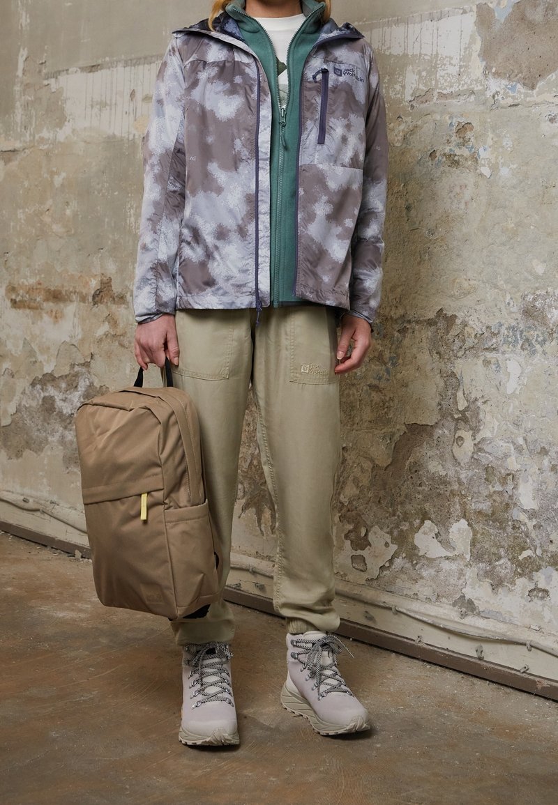 Lichte, waterdichte jas in het grijs met witte patronen, gelaagd over een groene ritssluiting, gecombineerd met khaki broek en een tan rugzak.
