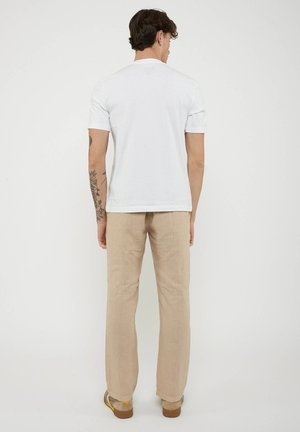 Man met een getatoeëerde arm, die een effen wit T-shirt met korte mouwen, beige broek en bruine schoenen draagt, staand vóór een witte achtergrond.