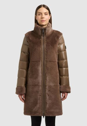 Une femme porte un long manteau marron avec un corps duveteux, des manches matelassées, un col montant et une fermeture éclair sur le devant.