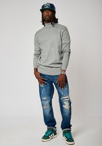 Suéter gris de cuello alto, jeans azules desgastados, zapatillas verdes y blancas, gorra de béisbol azul marino con texto. El modelo está de pie con una postura relajada.