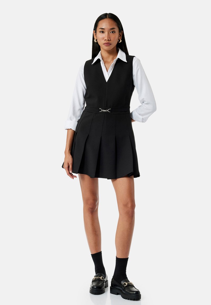 Zwarte jurk met een V-hals en een geplooide rok, gelaagd over een wit overhemd met knopen. Voorzien van een riem met een zilveren accent. Zwarte sokken en schoenen.