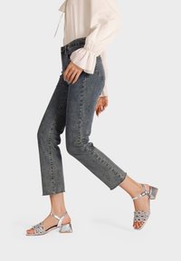 Sandali con tacco d'argento con anelli decorativi sulla parte superiore, sorretti da un tacco spesso. Abbinati a jeans in denim a vita alta e una camicia bianca.