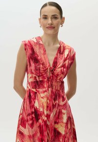 Sukienka maxi w intensywnym red i żółtym abstrakcyjnym wzorze. Ma dekolt w kształcie litery V z zapięciem na kokardę, krótkie rękawy i rozkloszowany dół.