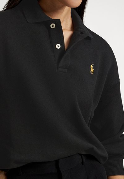 Črna polo majica iz mehke tkanine s trojnim gumbnim zapenjanjem, z majhnim zlatim grbom na levi strani prsi in rebrastimi manšetami.