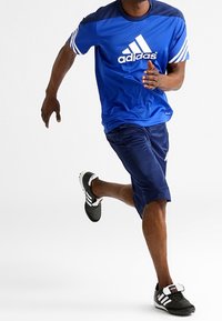 Sininen lyhythihainen urheilu paita, jossa on valkoinen Adidas-logo, yhdistettynä tummansinisiin sortseihin ja mustiin lenkkikenkiin, joissa on valkoiset raidat ja hihnakiinnitys.