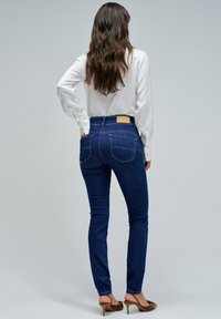 Jeans de denim azul ajustados con bolsillos traseros y una etiqueta marrón en la cintura, combinados con una blusa blanca de manga larga y zapatos de tacón marrones.