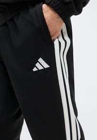 Χέρι στην τσέπη μαύρου παντελονιού Adidas με λευκό σχέδιο τριών ρίγων και λογότυπο Adidas στο μηρό.