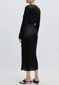 Schwarzes, plissiertes Midi-Kleid mit langen Ärmeln, tailliertem Schnitt, glatter Textur und Reißverschluss hinten, kombiniert mit schwarzen offenen High Heels.