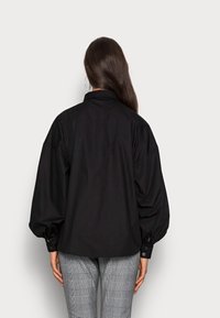 Black oversized shirt with a high collar, billowed sleeves, and button cuffs, made from smooth fabric. Paired with gray plaid trousers.