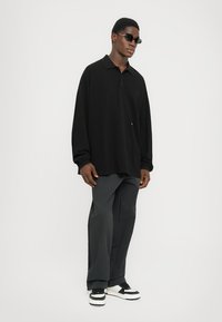 Schwarz langärmliges Poloshirt mit Kragen, kombiniert mit dunklen Hosen und schwarz-weißen Sneakers. Markante, eckige Sonnenbrille getragen.