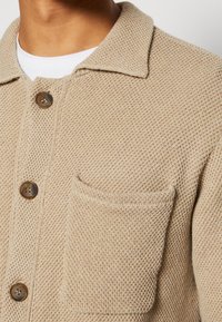 Chaqueta de punto texturizada en beige con cuello, botones frontales y un bolsillo en el pecho. Presenta un patrón tejido y detalles en botones marrones.