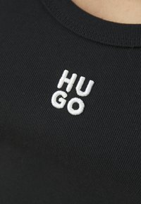 Zbliżenie na czarną żeberkowaną tkaninę z białymi haftowanymi literami "HUGO", podzielonymi na dwie linie w pobliżu dekoltu.