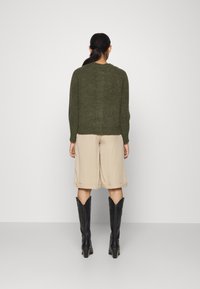 Olivgrön stickad tröja, beige shorts och svarta cowboyboots. Tröjan har långa ärmar och en rundad halsringning. Släta texturer synliga.