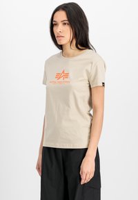 Béžové bavlněné tričko s oranžovým logem Alpha Industries, krátké rukávy, kulatý výstřih a malá černá visačka na rukávu.