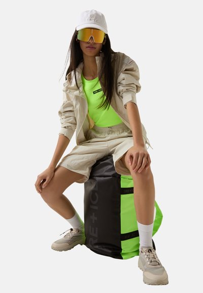Grøn og sort taske med fremtrædende logo, kombineret med en neongrøn top, beige jakke og shorts samt reflekterende solbriller. Afslappet atletisk look.